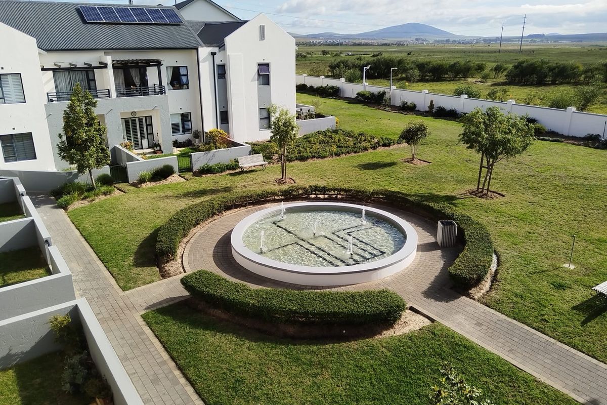 An elevated view of a well-maintained retirement village garden featuring a central circular fountain, paved walkways, and landscaped lawns with surrounding residential units.