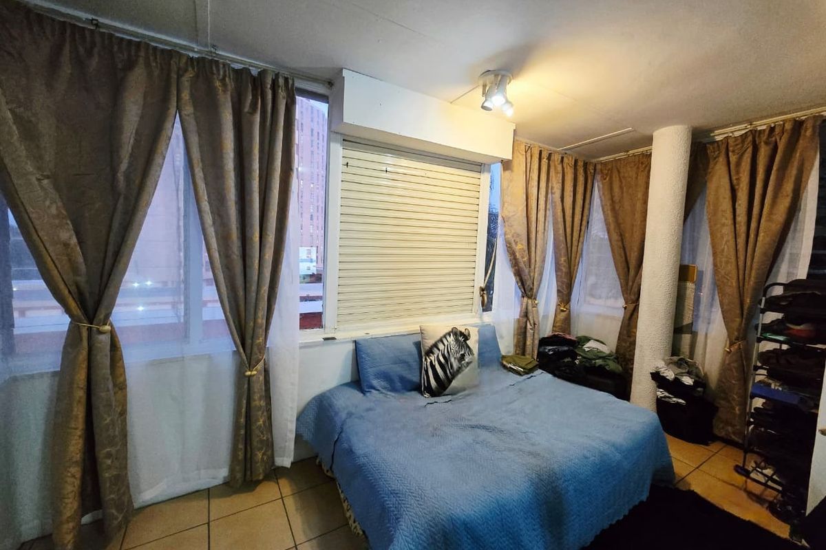 A bedroom featuring tiled flooring, a structural column, and a roller shutter window covering, providing a functional space with natural light.