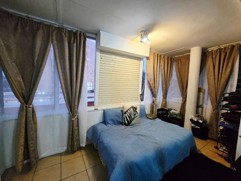 A bedroom featuring tiled flooring, a structural column, and a roller shutter window covering, providing a functional space with natural light.