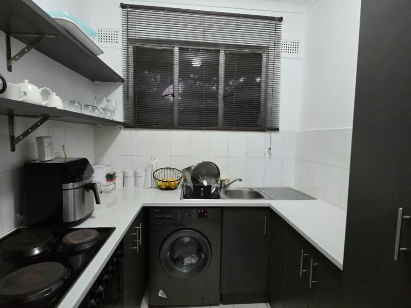 Compact kitchen featuring dark cabinetry, white countertops, tiled splashback, integrated electric cooktop, and tiled flooring with a window for natural light.