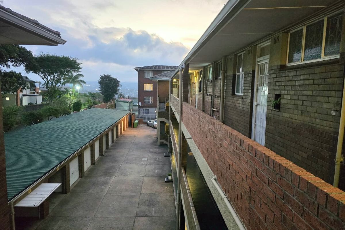 An elevated view of an apartment complex exterior, featuring a brick facade, communal walkway, and a row of garages in the courtyard.