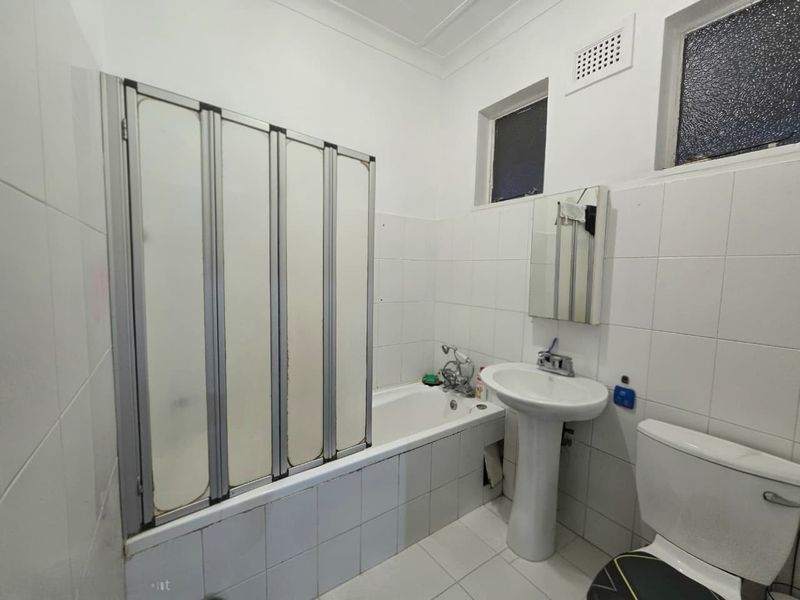 A functional bathroom featuring a pedestal basin, toilet, and a bathtub with a folding shower screen, finished with white wall and floor tiles.