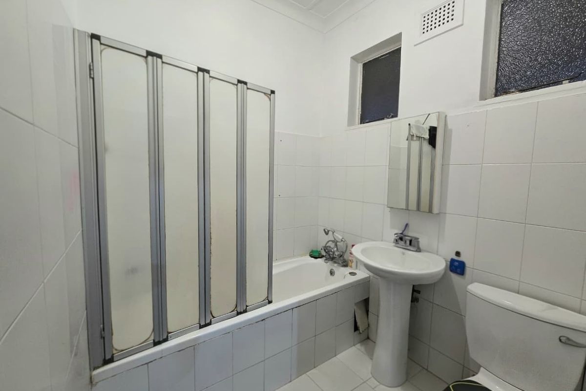 A functional bathroom featuring a pedestal basin, toilet, and a bathtub with a folding shower screen, finished with white wall and floor tiles.