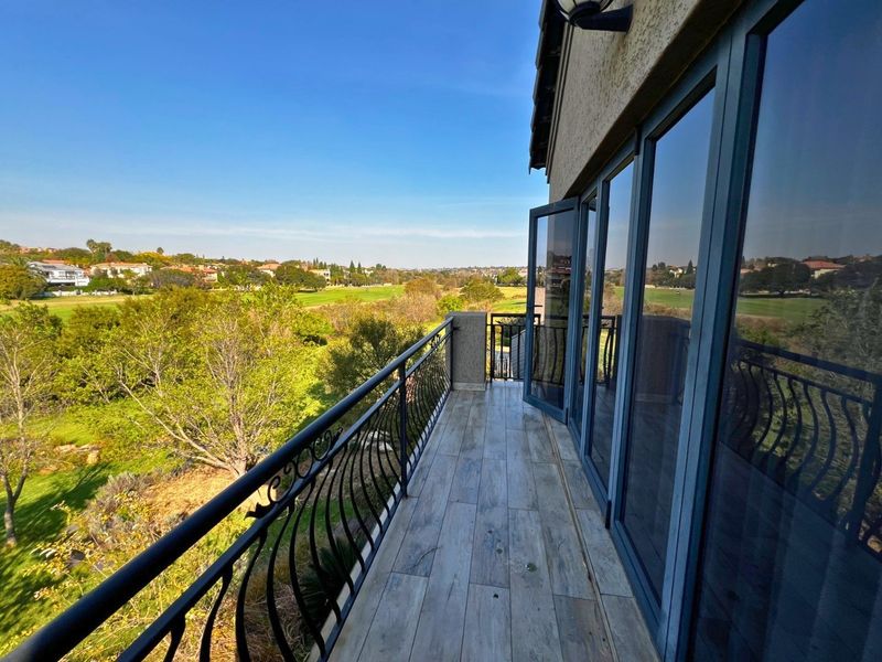 A spacious balcony featuring wood-look tiled flooring, decorative wrought iron railings, and expansive glass folding doors overlooking a scenic golf course landscape.