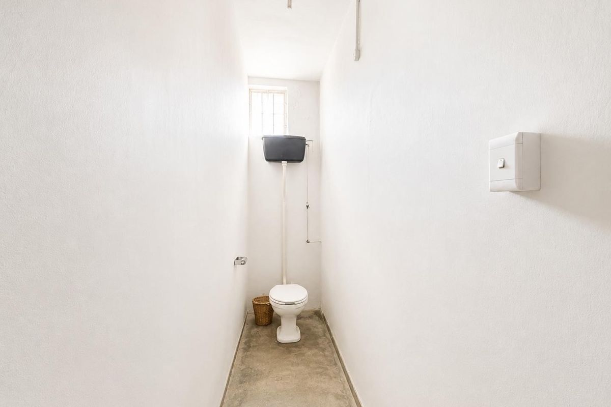 A compact, utilitarian restroom featuring a high-level cistern toilet, exposed surface-mounted electrical conduit, and a simple concrete floor finish.
