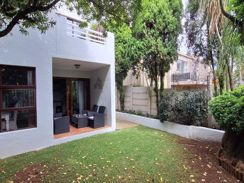 A rear view of the property featuring a covered patio, an upper-level balcony, and a private garden with established trees and boundary walling.