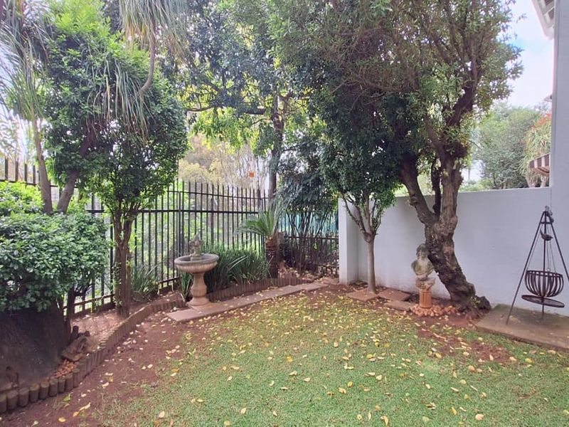 A private garden area featuring mature trees, a stone bird bath, and secure metal perimeter fencing, adjacent to a paved patio section.