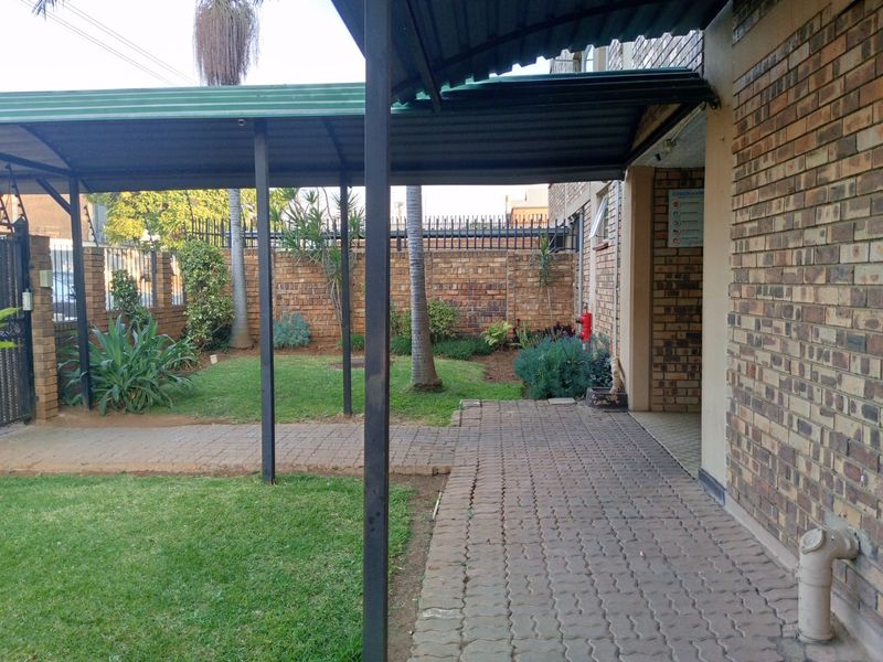 A covered walkway with paved path leading through a garden area, featuring brick boundary walls and metal roofing structure.