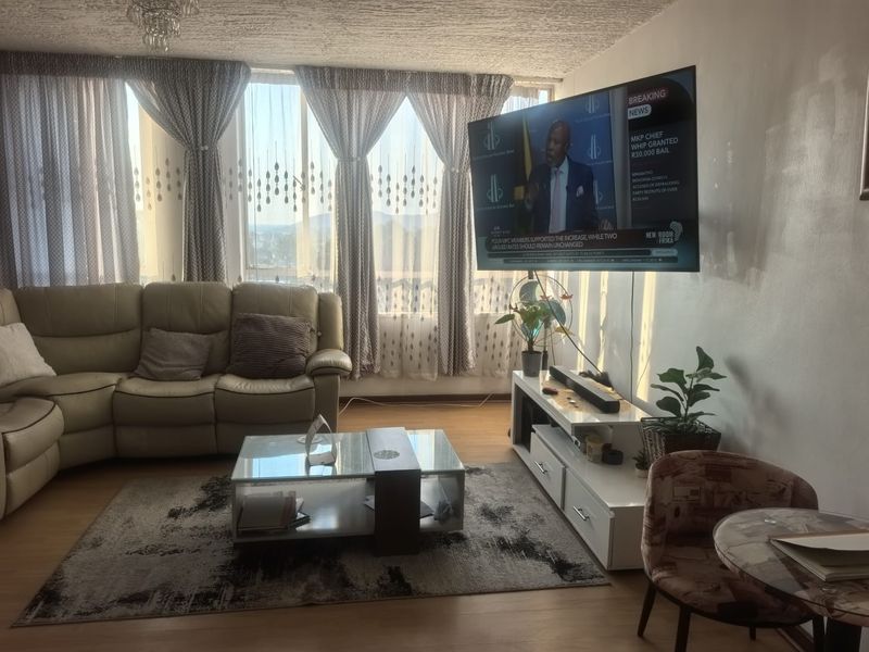 A living room featuring large windows with natural light, laminate flooring, and a wall-mounted television.