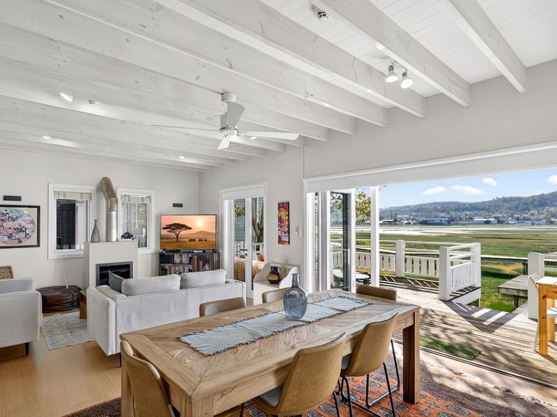 Open-plan living and dining area featuring exposed white timber beams, a built-in fireplace, and large folding doors opening onto a wooden deck with views.