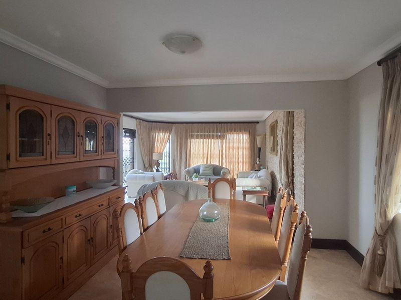 A spacious open-plan dining and living area featuring neutral wall tones, tiled flooring, and large windows with fitted blinds.