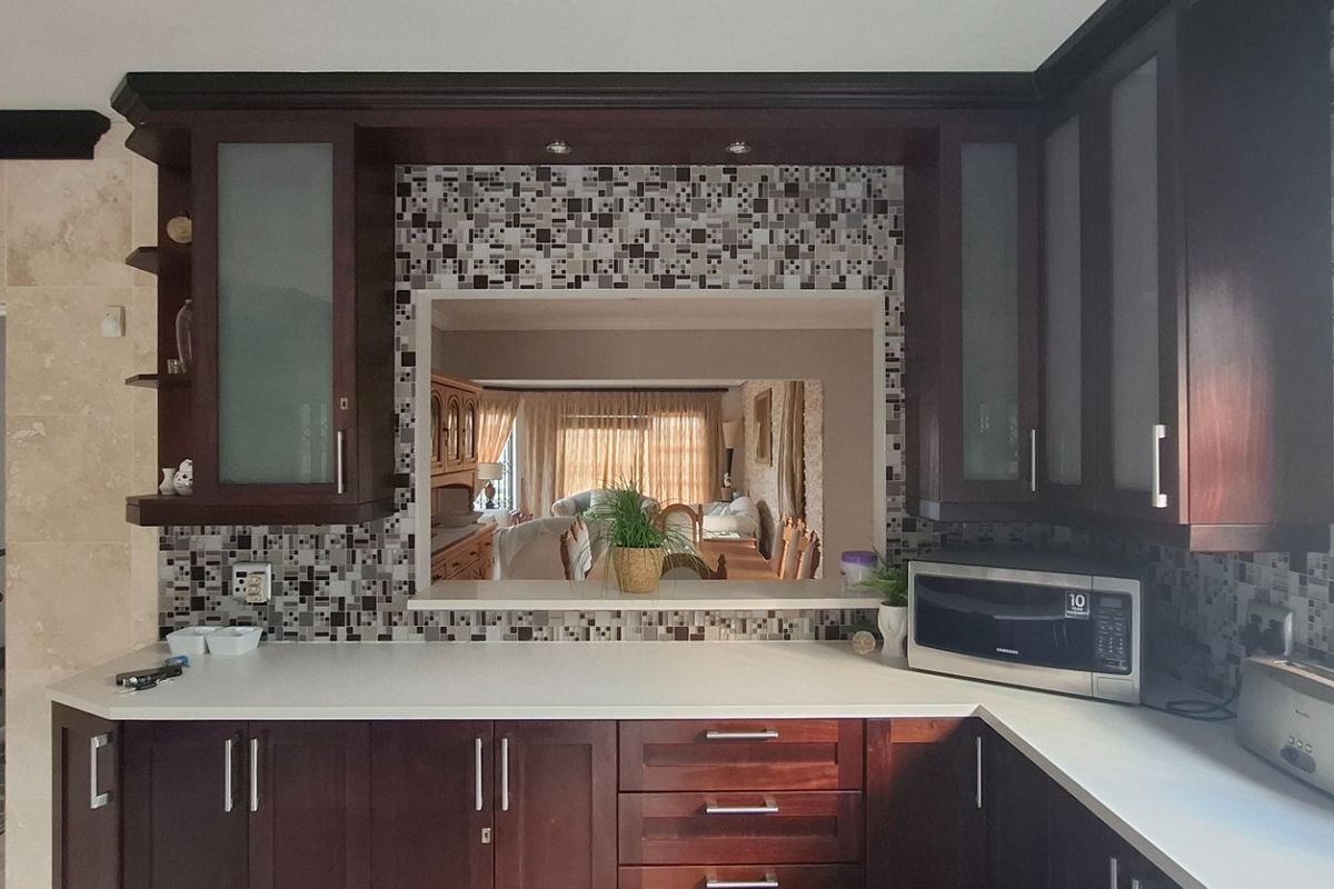 A kitchen featuring dark wood cabinetry, mosaic tile backsplash, and a serving hatch looking through to the adjacent living area.