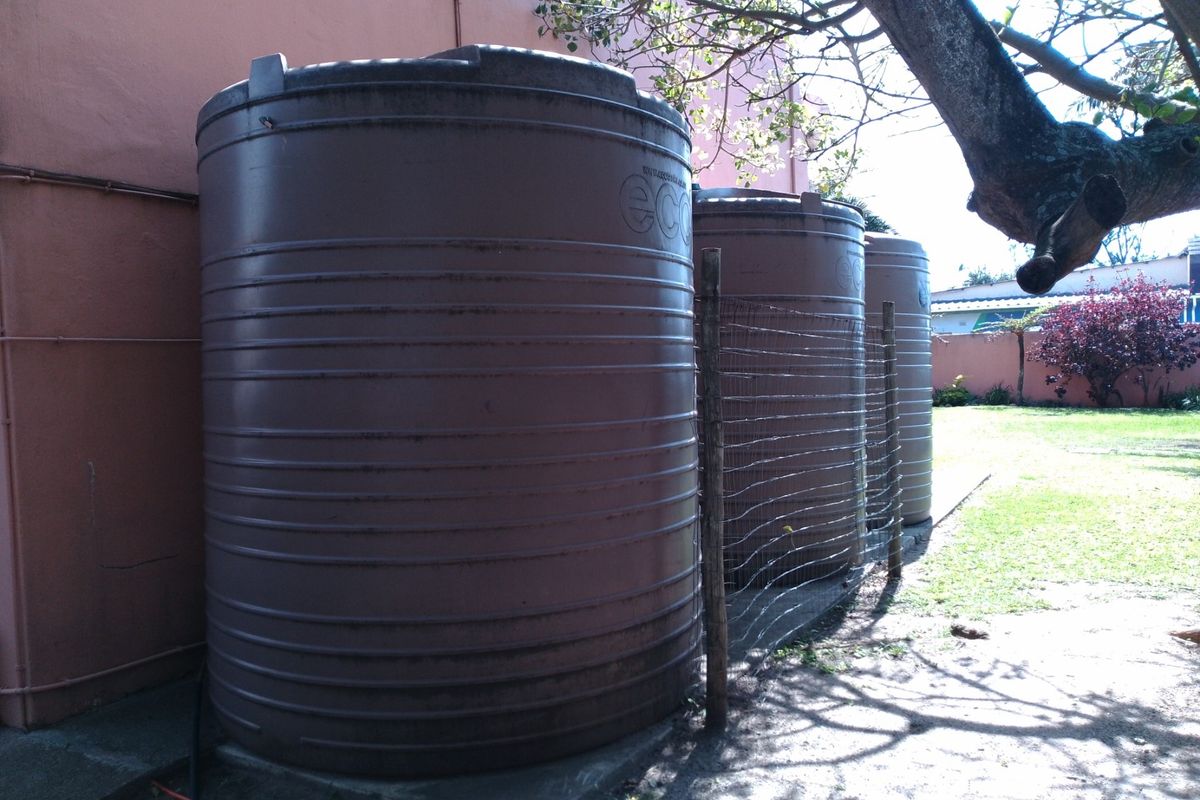 Exterior view of three large vertical water storage tanks positioned against the side of the house, situated on a concrete base in the garden.