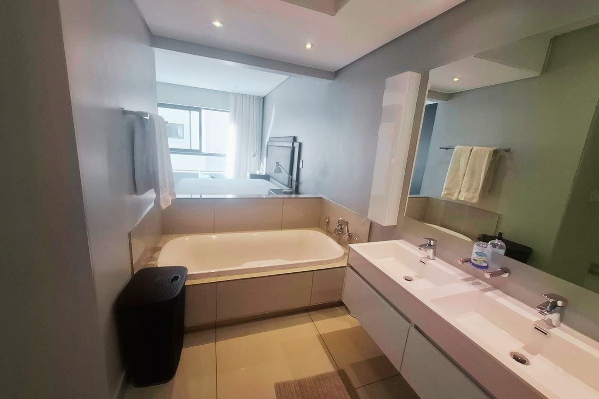 Modern en-suite bathroom featuring a built-in bathtub, double vanity with integrated sinks, and large wall-mounted mirror, with an open-plan view into the bedroom.