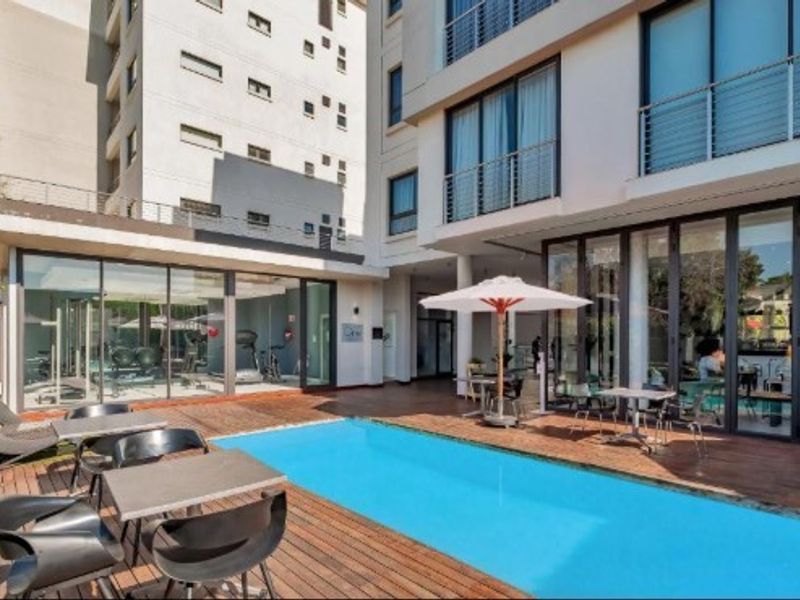 A well-maintained apartment complex pool area featuring a swimming pool, wooden decking, and adjacent glass-enclosed gym facility.