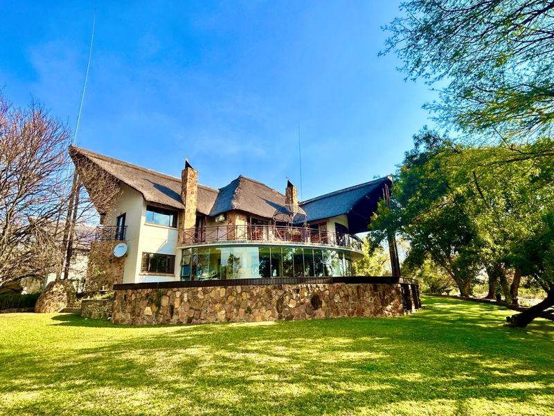 Rear view of a unique architectural home featuring a thatched roof, stone cladding, and a prominent curved glass wall overlooking a spacious garden.