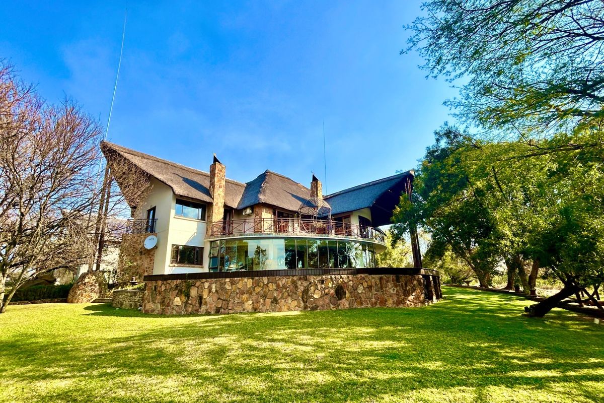 Rear view of a unique architectural home featuring a thatched roof, stone cladding, and a prominent curved glass wall overlooking a spacious garden.