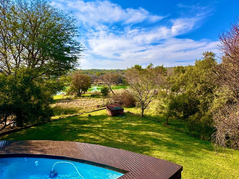 An elevated view of a swimming pool with a wooden deck overlooking a spacious, tree-lined garden and natural landscape.