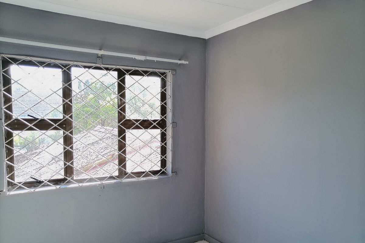 A bright, empty bedroom featuring grey painted walls, tiled flooring, and a window fitted with security burglar bars.