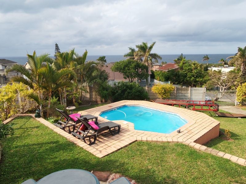An elevated view of a backyard featuring a swimming pool with brick coping, surrounded by a landscaped garden and perimeter fencing with ocean views.