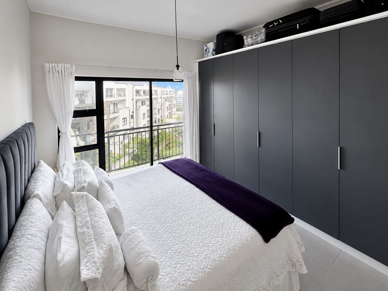A well-maintained bedroom featuring floor-to-ceiling built-in cupboards, large windows with balcony access, and neutral tiled flooring.