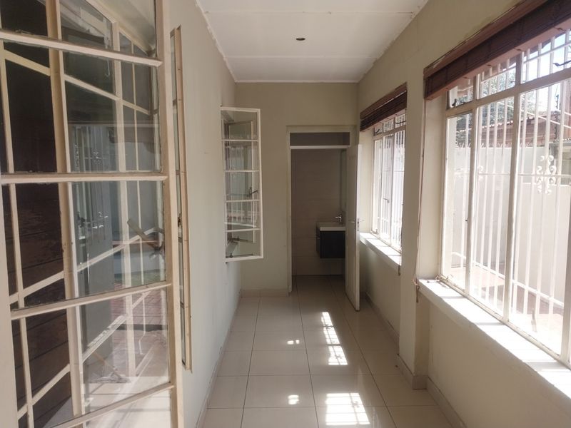 A bright, tiled hallway featuring large windows with security bars, leading towards a small bathroom area.