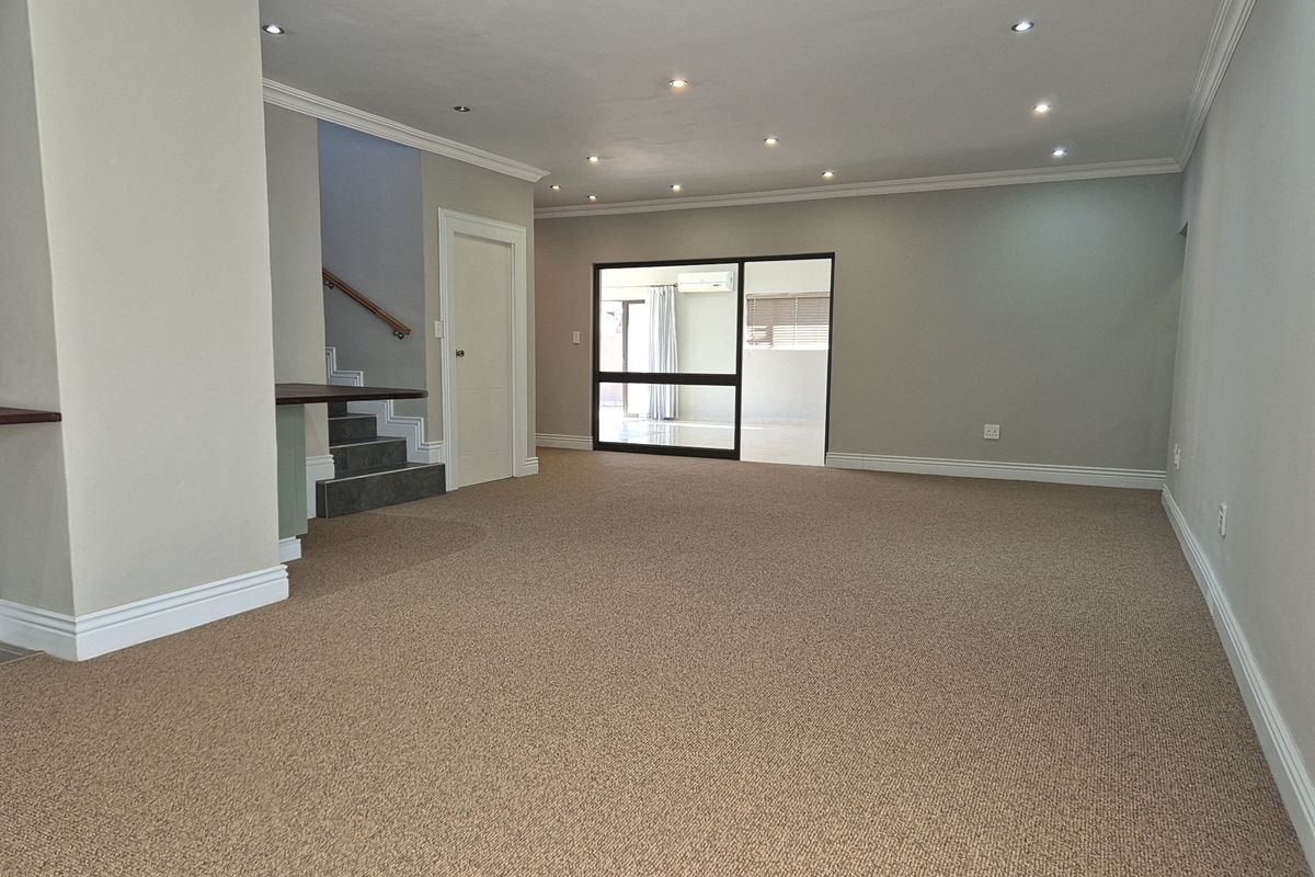 A spacious, empty living area featuring neutral carpeting, recessed ceiling lighting, crown molding, and a staircase leading to the upper level.