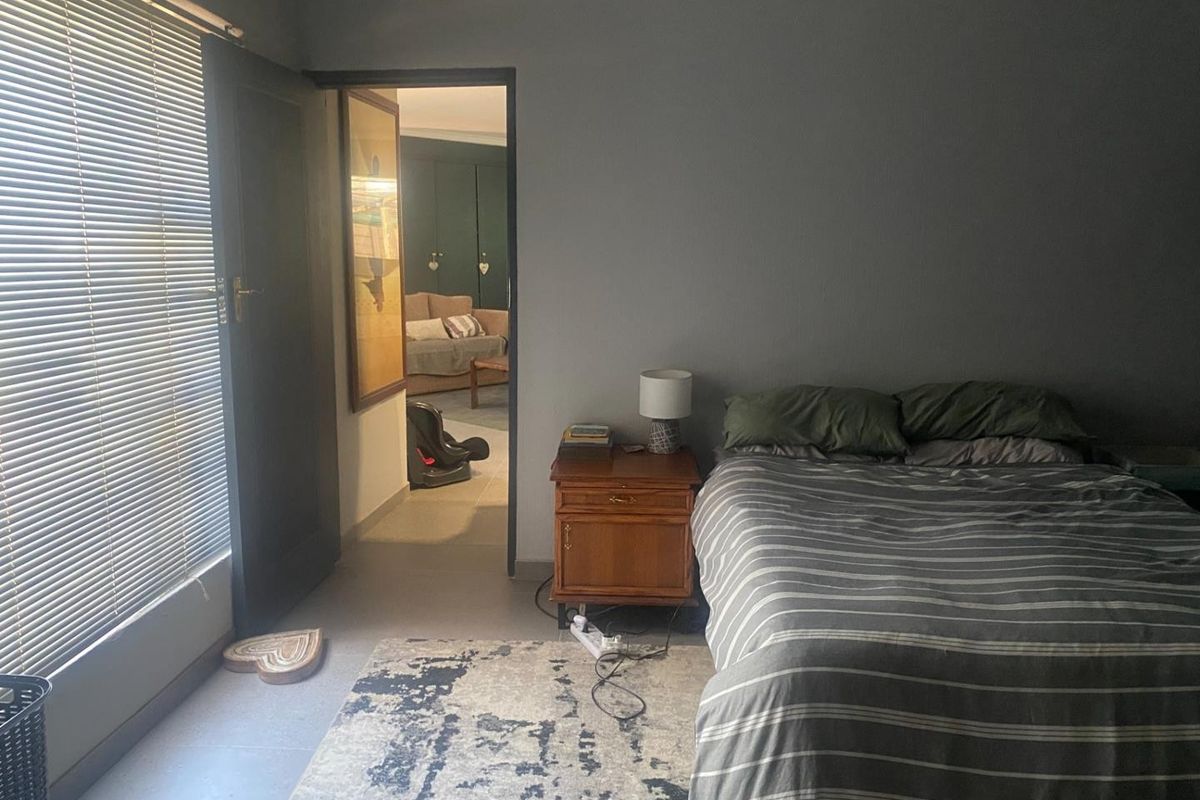 A bedroom featuring grey walls, tiled flooring, and venetian blinds, with a doorway leading to an adjacent living space.