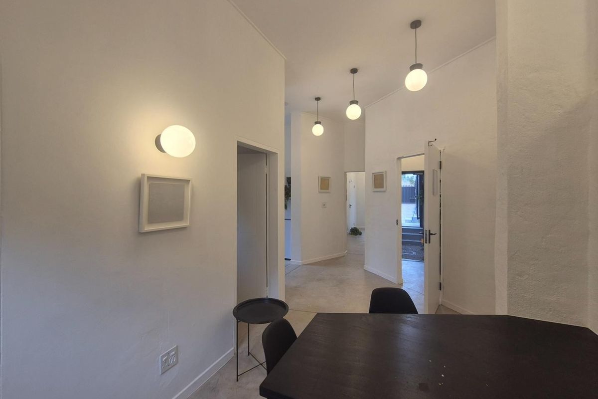 A bright, minimalist hallway featuring modern pendant lighting, wall-mounted sconces, and clean white finishes, providing access to multiple rooms within the commercial space.