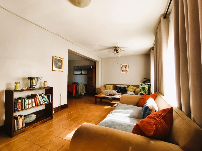 A spacious open-plan living area featuring terracotta floor tiles, a ceiling fan, and large windows allowing for ample natural light.