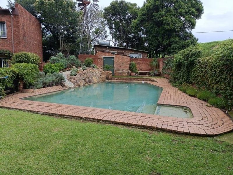 A private backyard area featuring a swimming pool with brick paving, surrounded by established garden beds and a brick boundary wall.