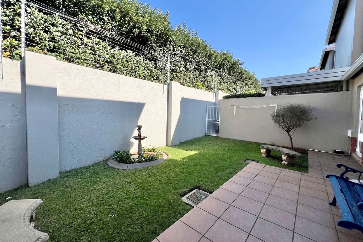 A private rear garden featuring a paved patio area, a small lawn, a decorative water feature, and high perimeter walls with electric fencing.