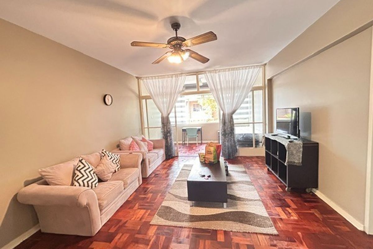 A spacious living room featuring classic parquet flooring, a ceiling fan, and large glass sliding doors leading to an enclosed balcony area.