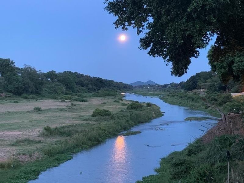 A scenic view of a river flowing through a natural landscape at dusk with the moon visible in the sky.