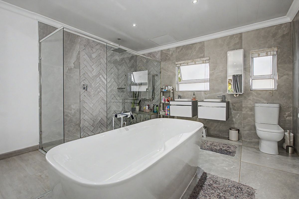 Modern bathroom featuring a freestanding bathtub, large glass-enclosed walk-in shower with herringbone tile detail, wall-mounted dual vanities, and contemporary grey floor-to-ceiling tiling.