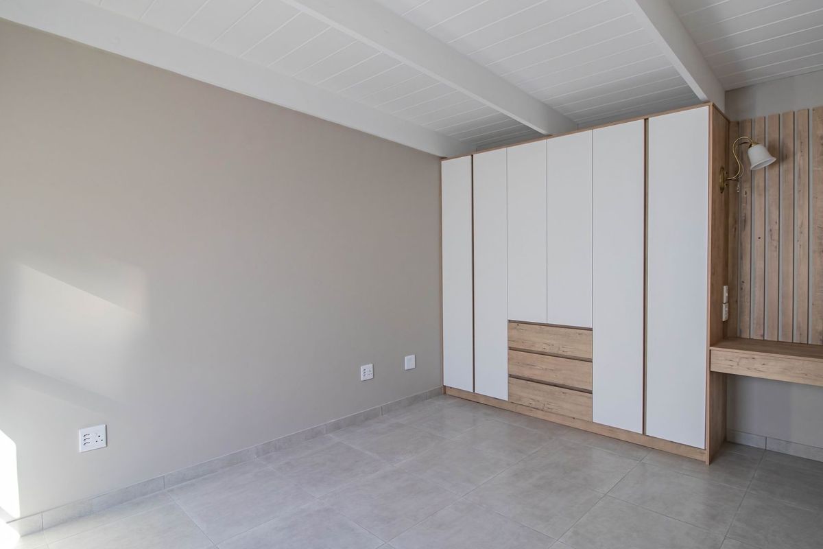 A bright, modern bedroom featuring neutral tones, tiled flooring, and a large, custom-built floor-to-ceiling wardrobe with an integrated vanity desk.