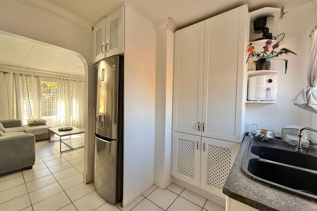 A functional kitchen area featuring white cabinetry, a double sink, and tiled flooring, seamlessly transitioning into an adjacent living space through an arched doorway.