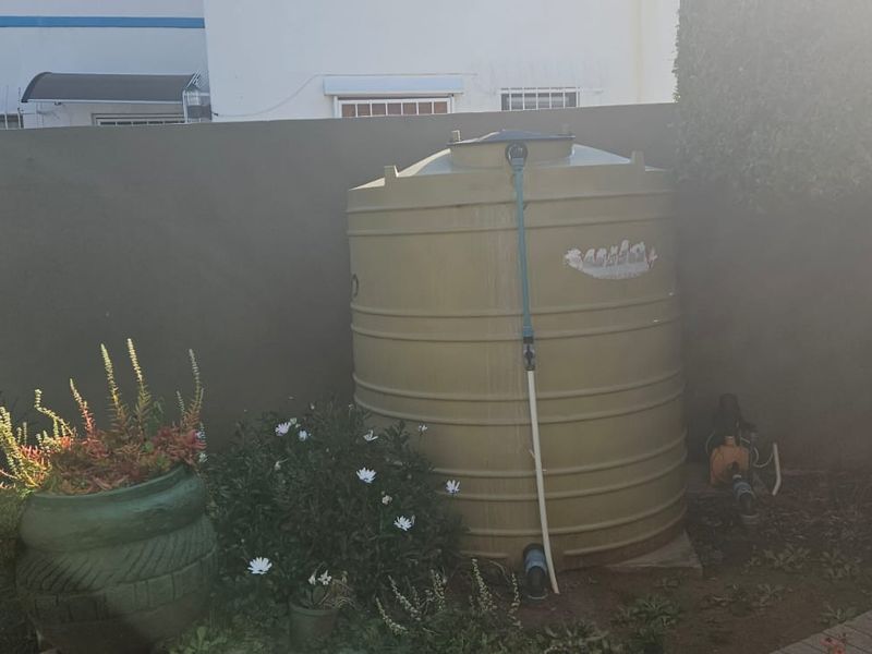 A residential garden area featuring a large plastic water storage tank and a connected water pump system against a boundary wall.