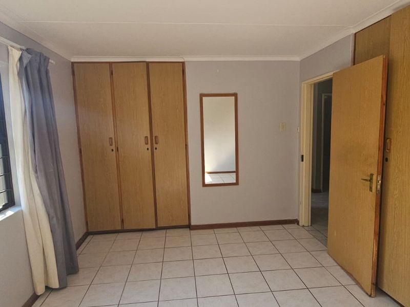 A bedroom featuring tiled flooring, built-in wooden cupboards, and a window with security bars overlooking the garden.