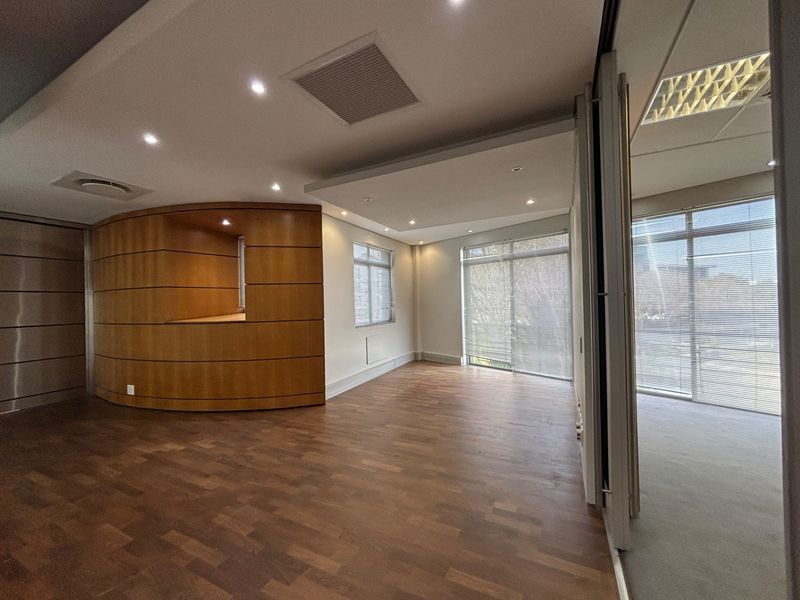 A modern, vacant office space featuring high-quality wooden parquet flooring, a distinctive curved wooden feature wall, recessed lighting, and large windows with integrated blinds.