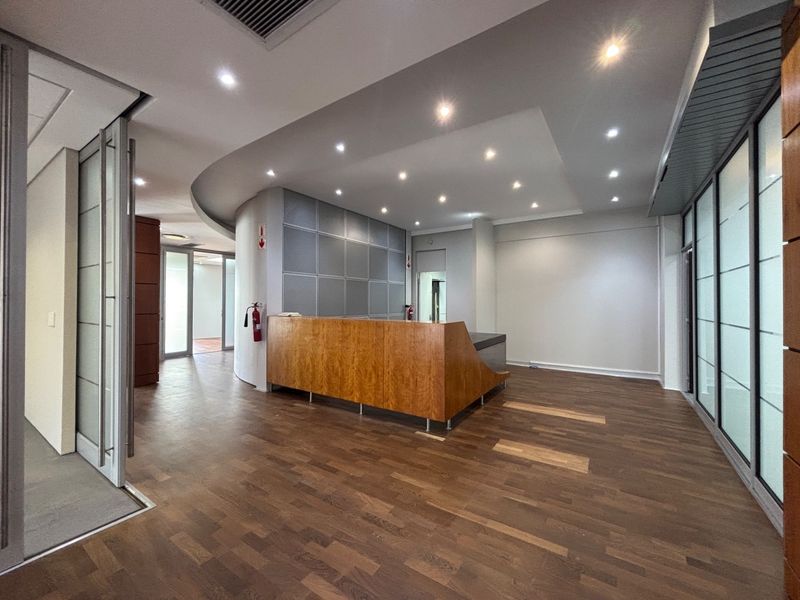 A professional office reception area featuring a built-in wooden desk, recessed ceiling lighting, and modern glass partitions with wood laminate flooring.