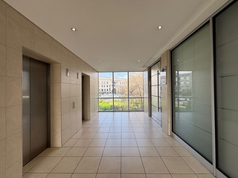 A bright and modern office building hallway featuring tiled flooring, elevator access, and large windows providing natural light and exterior views.