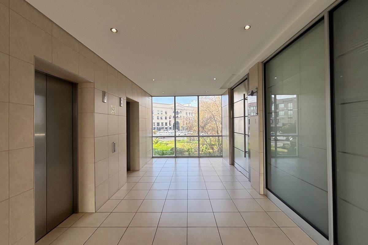 A bright and modern office building hallway featuring tiled flooring, elevator access, and large windows providing natural light and exterior views.