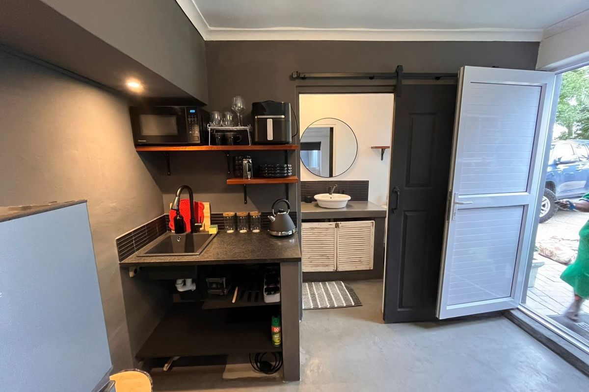 A compact kitchenette featuring a dark countertop, integrated sink, and open shelving, with an adjacent bathroom accessible via a sliding barn door.
