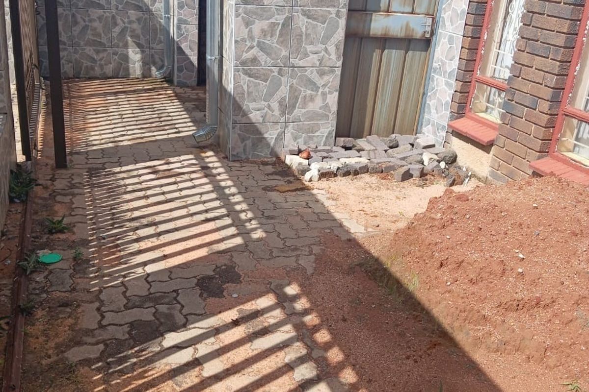 A side exterior area featuring paved sections, tiled wall cladding, and an access door, currently requiring landscaping and maintenance to complete the outdoor space.