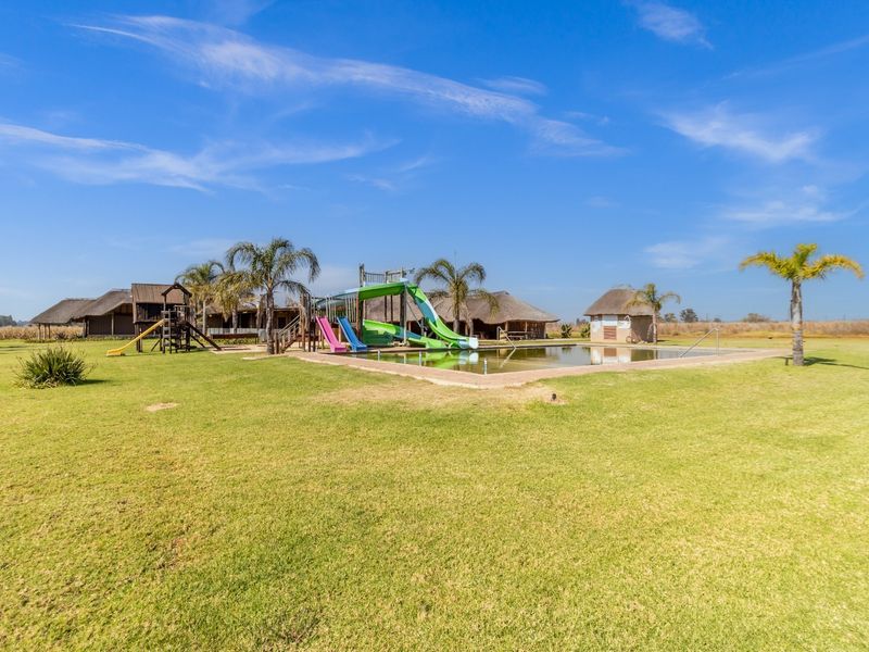 Expansive farm grounds featuring a swimming pool with integrated slides, a children's play structure, and traditional thatched-roof buildings in a rural setting.
