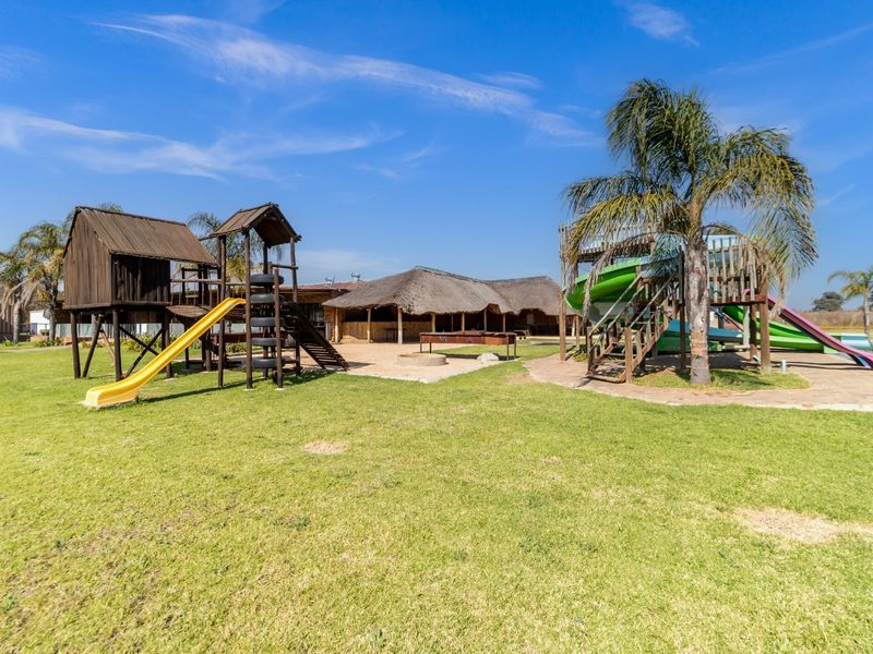 Expansive outdoor recreational area featuring multiple wooden jungle gyms, slides, and a thatched-roof entertainment structure set within a large, open garden space.