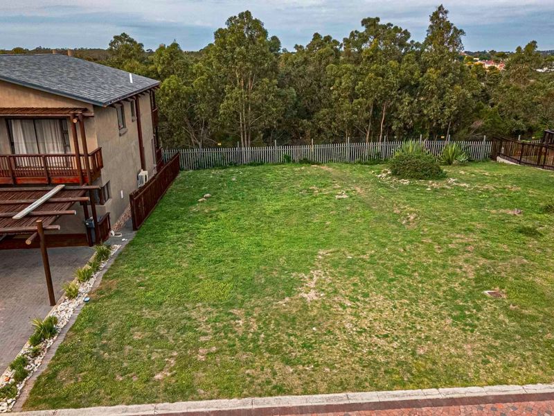 An elevated view of a residential backyard featuring a grassed area, paved driveway section, and perimeter fencing, adjacent to a multi-story dwelling.