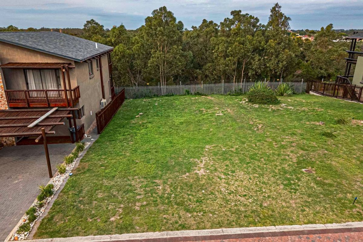 An elevated view of a residential backyard featuring a grassed area, paved driveway section, and perimeter fencing, adjacent to a multi-story dwelling.
