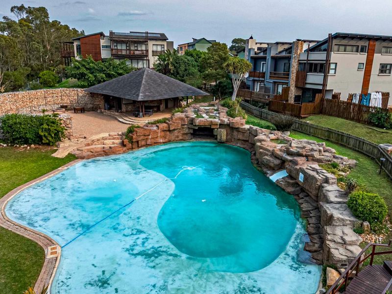 Aerial view of a landscaped residential pool area featuring a rock-feature swimming pool, stone retaining wall, and a covered outdoor entertainment pavilion.